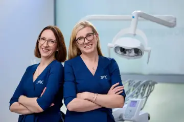 Dwie uśmiechnięte kobiety w granatowych uniformach medycznych, w gabinecie stomatologicznym.