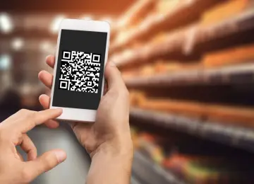 Dłoń trzyma smartfon z widoczną na ekranie aplikacją do skanowania kodów QR. Tło sugeruje sklep.