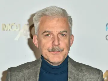 Hubert Urbański z siwymi włosami i wąsami, w szarym garniturze i granatowym golfie. W tle logo "Mój".