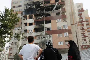 Zniszczony budynek mieszkalny, ludzie patrzą na pożar, w tle drabina strażacka.
