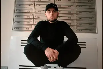Bedoes w czapce z daszkiem i czarnym ubraniu kuca przed skrzynkami na listy. Jego wzrost jest typowy dla mężczyzny.