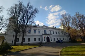 Pałac w Falentach, biała, okazała budowla z wieżyczką, otoczona drzewami o nagich gałęziach, pod błękitnym niebem z chmurami.