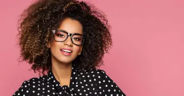 Une jeune femme aux cheveux bouclés porte un maquillage éclatant et des lunettes noires, souriant sur fond rose.