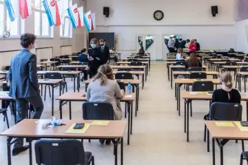 Uczniowie siedzą w ławkach podczas egzaminu. W tle widać flagi, co może sugerować, jakie są rozszerzenia w liceum.