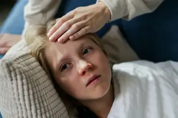 Dziecko z gorączką leży na poduszce, a ręka mamy sprawdza mu czoło. Czy dziecko z gorączką może wychodzić na dwór?