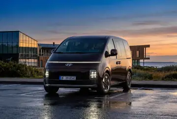 Ciemnobrązowy Hyundai Staria van zaparkowany na mokrym parkingu o zachodzie słońca.