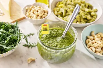 Pesto z rukoli w słoiku, obok miseczki z rukolą, orzechami nerkowca i makaronem. Idealne do letnich dań.