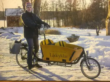 Man med mössa och jacka cyklar på vintern med en gul lastcykel i snöig miljö.