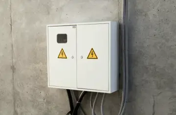 Biała skrzynka elektryczna w betonowym ogrodzeniu, z żółtymi symbolami ostrzegawczymi.