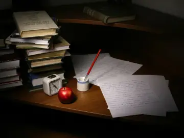 Stos książek, jabłko, budzik i pióro z kałamarzem na biurku. Inspiracja do napisania felietonu.