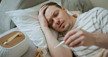 Chory mężczyzna leży w łóżku, trzymając termometr. Wygląda na to, że zmaga się z objawami grypy typu K.