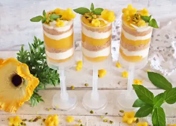 Warstwowy deser z mango w przezroczystych pojemnikach, ozdobiony listkami mięty i pistacjami.
