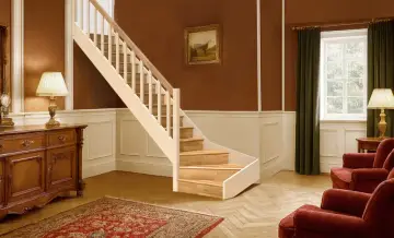 Eleganckie schody w stylu angielskim, z drewnianymi stopniami i jasną balustradą, prowadzą do przytulnego wnętrza z kominkiem i obrazem na ścianie.