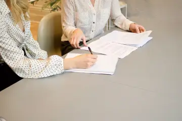 Dwie kobiety przy stole omawiają umowę przedwstępną sprzedaży nieruchomości. Jedna pisze notatki.