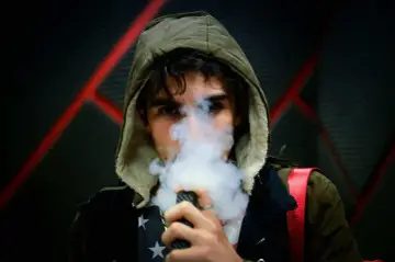 Joven con capucha exhala vapor de un vapeador, con un diseño de estrellas en su camiseta. Los nicokits son su secreto.