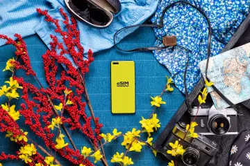 Żółty telefon z napisem eSIM obok mapy, aparatów i letnich ubrań. Idealny na wakacje w Turcji, bez obaw o roaming.
