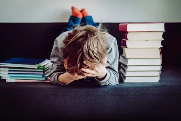 Chłopiec z rękami na twarzy, przytłoczony stertą książek. Może to znak, że potrzebne jest orzeczenie z poradni psychologiczno-pedagogicznej.