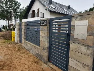 Nowoczesne ogrodzenie z ciemnoszarych paneli i słupków z kamienia, z widocznym numerem posesji.