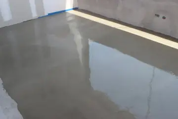 Świeżo wylany beton na posadzkę, błyszczący od wilgoci, czeka na stwardnienie. W tle widać ściany i niebieską taśmę.