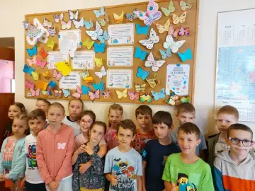 Dzieci z uśmiechem na twarzy pozują przed tablicą pełną kolorowych motyli i cytatów o dobrych uczynkach.