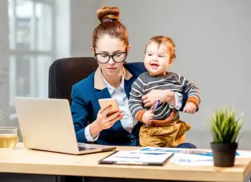 Pracująca mama z dzieckiem na rękach sprawdza telefon. Kto wypłaca macierzyński pracodawca czy ZUS?