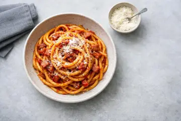 En skål med rörig pasta amatriciana, toppad med riven ost. En liten skål med mer ost och en grå servett syns bredvid.