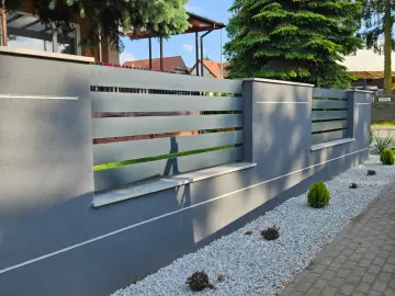 Nowoczesny płot beton architektoniczny z szarymi, poziomymi przęsłami. Ozdobne kamyczki i miniaturowe krzewy tworzą schludny ogród.