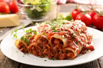 Pyszne cannelloni z mięsem mielonym, polane sosem pomidorowym i posypane parmezanem, na tle świeżych pomidorów i sałatki.