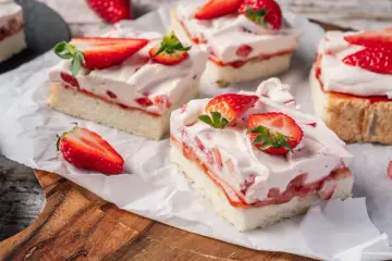 Saftige Stücke eines Erdbeerkuchen Blechs mit frischen Erdbeeren und Sahne, auf Backpapier angerichtet.