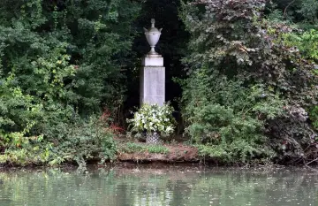 Kamienny pomnik z urną, otoczony zielenią, z bukietem białych kwiatów u podstawy. Miejsce pamięci, przypominające grób księżnej Diany.