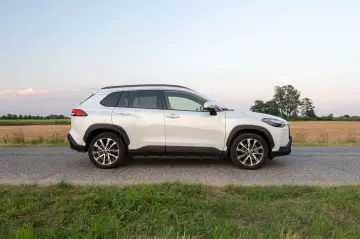 Biała Toyota Corolla Cross Hybrid stoi na poboczu drogi, otoczona polami i zieloną trawą.