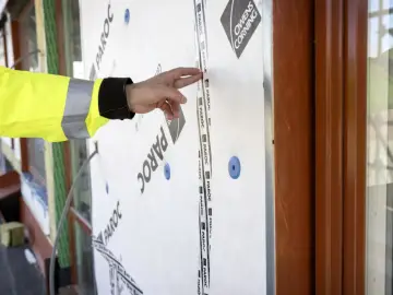 Ręka w żółtej kamizelce wskazuje na taśmę na materiale budowlanym. To kluczowy element wiatroizolacji, chroniący budynek przed wiatrem i wilgocią.