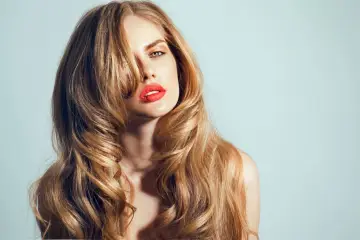 Kobieta z burzą falujących, blond włosów, ułożonych w modny butterfly cut. Jej usta podkreślone są jaskrawą czerwienią.