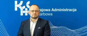 Naczelnik urzędu skarbowego w garniturze, na tle niebieskiej ściany z logo Krajowej Administracji Skarbowej.