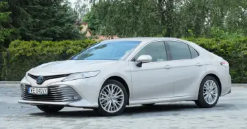 Srebrna Toyota Camry 2.5 Hybrid stoi na wybrukowanym podjeździe, otoczona zielenią.