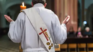 Ksiądz w białej szacie z krzyżem i kłosem na stułę, z rękami wzniesionymi w geście modlitwy, tworzy atmosferę religijnej suspensy.