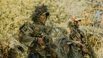 Dwóch snajperów w ghillie suit, ukrytych w zaroślach, czeka na cel.