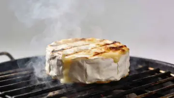 Un camembert au barbecue, fondant et fumant, cuit sur une grille. La croûte blanche se craquelle, révélant un cœur doré et coulant.