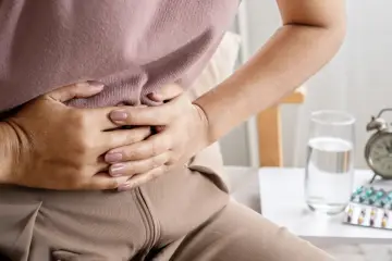 Kobieta trzyma się za brzuch, cierpiąc z powodu wzdęć. Obok stoi szklanka wody i opakowania leków.