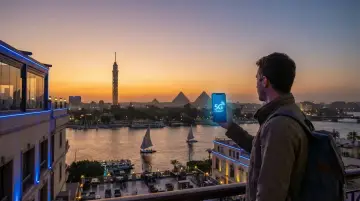 Mężczyzna z plecakiem trzyma telefon z napisem "5G CONNECT" na tle zachodzącego słońca nad Nilem, z widocznymi piramidami w Egipcie. eSIM Egipt zapewnia łączność.