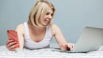 Kobieta z telefonem w ręku uczy się, jak udostępnić internet z laptopa, leżąc w łóżku.