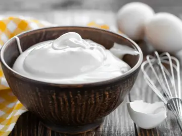 Un bol rempli de crème fouettée, blanc en neige, prêt à être utilisé pour une pâtisserie. Des œufs et un fouet sont visibles à proximité.