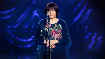 Grażyna Łobaszewska w kwiecistej sukni, gotowa zaśpiewać swój przebój na tle niebieskich świateł.
