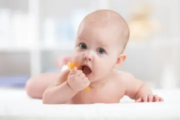 Niemowlę na brzuchu gryzie pomarańczową zabawkę. Czasem późne ząbkowanie przyczyny ma genetyczne.