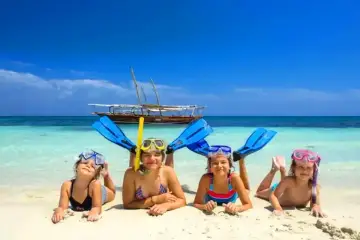 Cztery uśmiechnięte dzieci w maskach do snorkelingu i płetwach leżą na plaży. W tle turkusowa woda i łódź. Idealny dzień, kiedy na Zanzibar!
