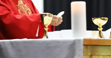 Kapłan w czerwonej szacie, trzymający hostię, przygotowuje się do komunii. Obok kielichy i świeca. Dyskusja o podatek na kościół trwa.