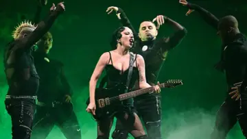 Lady Gaga koncert, w czarnym stroju z gitarą, śpiewa na scenie otoczona tancerzami w podobnych stylizacjach.