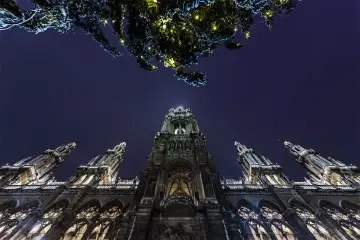 Majestatyczny budynek w stylu neogotyckim nocą, oświetlony, z widocznym fragmentem choinki z lampkami.