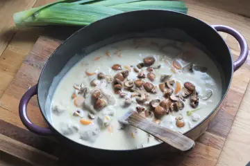 Un plat de blanquette de veau facile mijote dans une cocotte, garni de champignons et de carottes. Un poireau est posé à côté.