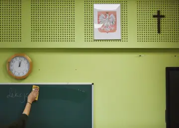 Klasa z zegarem, godłem Polski i krzyżem. Ręka zmywa napis z tablicy, jakby to był wzór rezygnacji ze szkoły podstawowej.